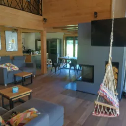 Jasne wnętrze domu z bali z drewnianymi ścianami, szarymi sofami, wiszącym fotelem hamakowym i nowoczesnym kominkiem z telewizorem