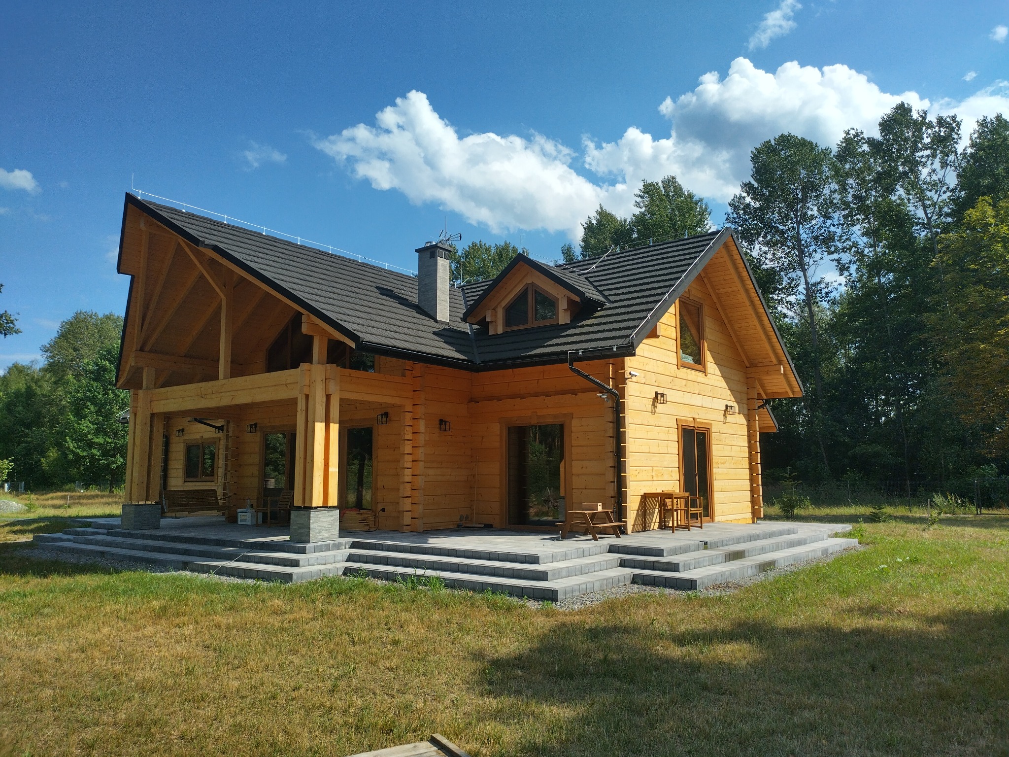 Nowoczesny, dwukondygnacyjny dom z bali z ciemną dachówką, widoczną konstrukcją drewnianą i kamiennymi schodami wejściowymi, otoczony zielenią i błękitnym niebem z chmurami.