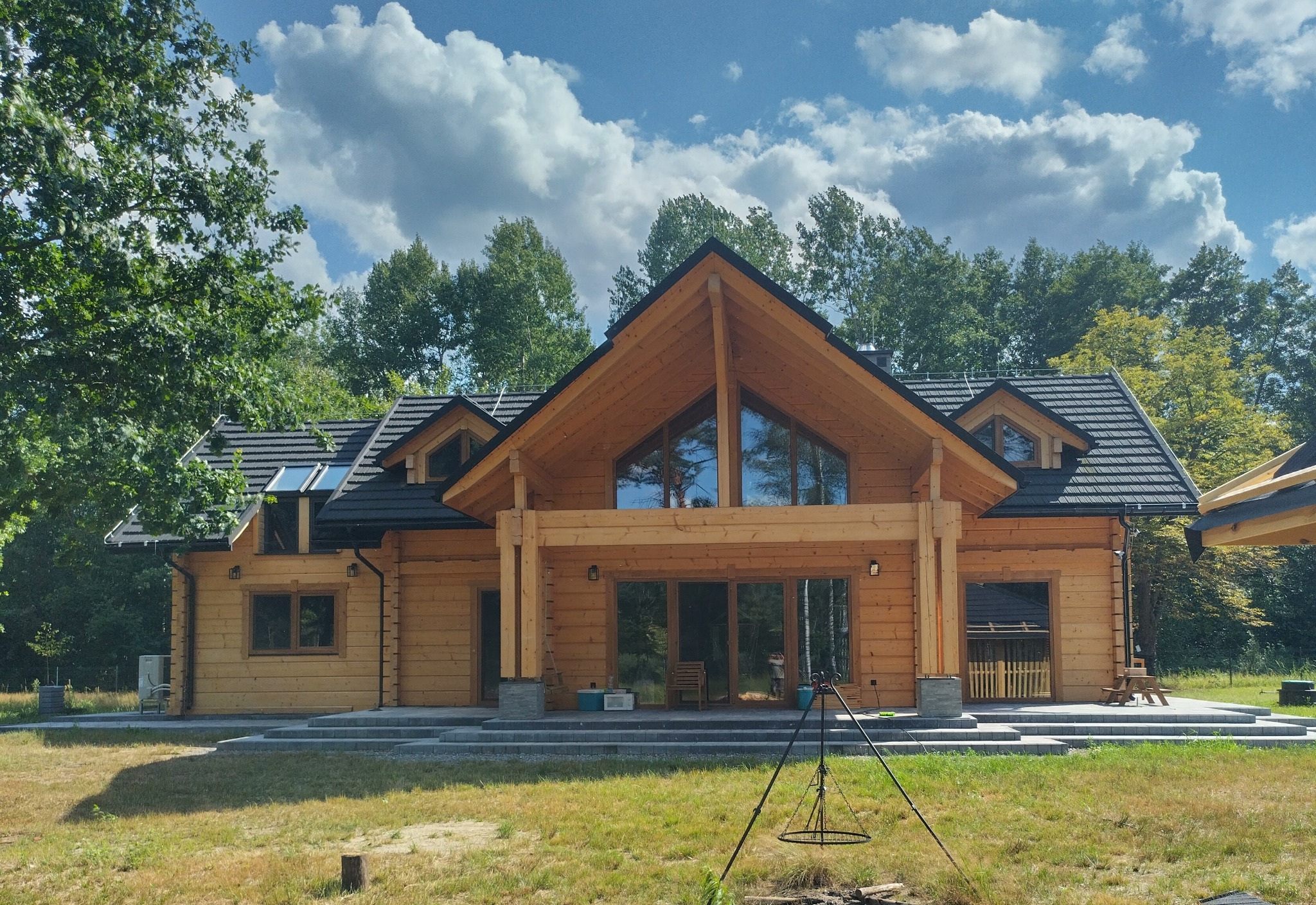 Gotowy, parterowy dom z bali z czarnym dachem i dużymi przeszkleniami, na tle zielonego lasu i błękitnego nieba z chmurami. Na pierwszym planie miejsce na ognisko.