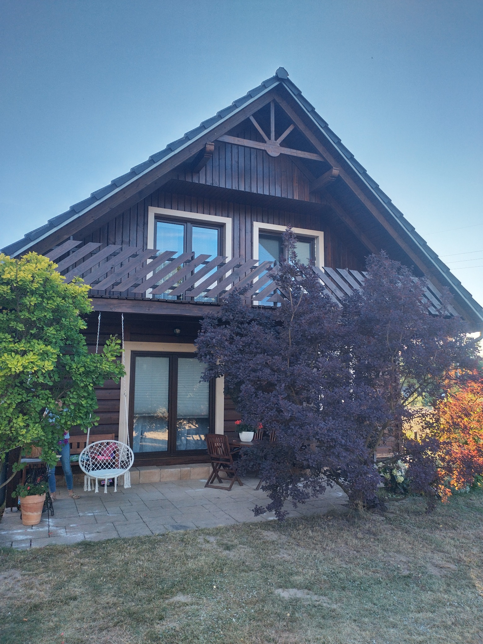 Brązowy dom z bali z balkonem i ciemną więźbą dachową, otoczony zielenią i fioletowym drzewem, z wiszącym krzesłem na tarasie.