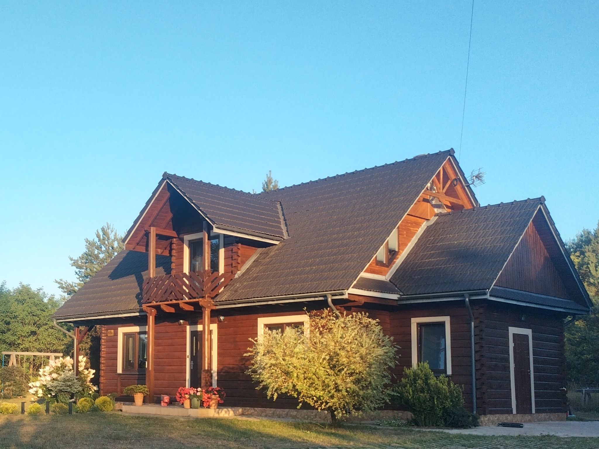 Ciemnobrązowy dom z bali z dwuspadowym dachem z ciemnej dachówki, z drewnianą balustradą balkonu, otoczony zielenią w słoneczny dzień.