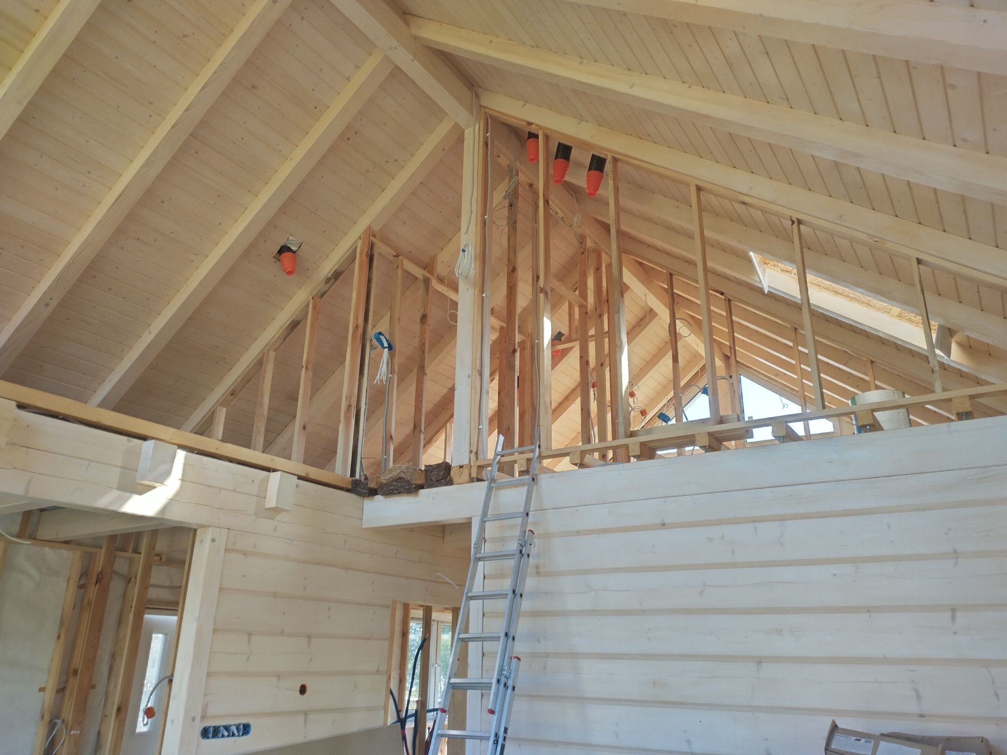 Wnętrze domu w trakcie budowy, widoczna drewniana konstrukcja ścian i dachu z elementami instalacji elektrycznej, drabina oparta o ścianę.