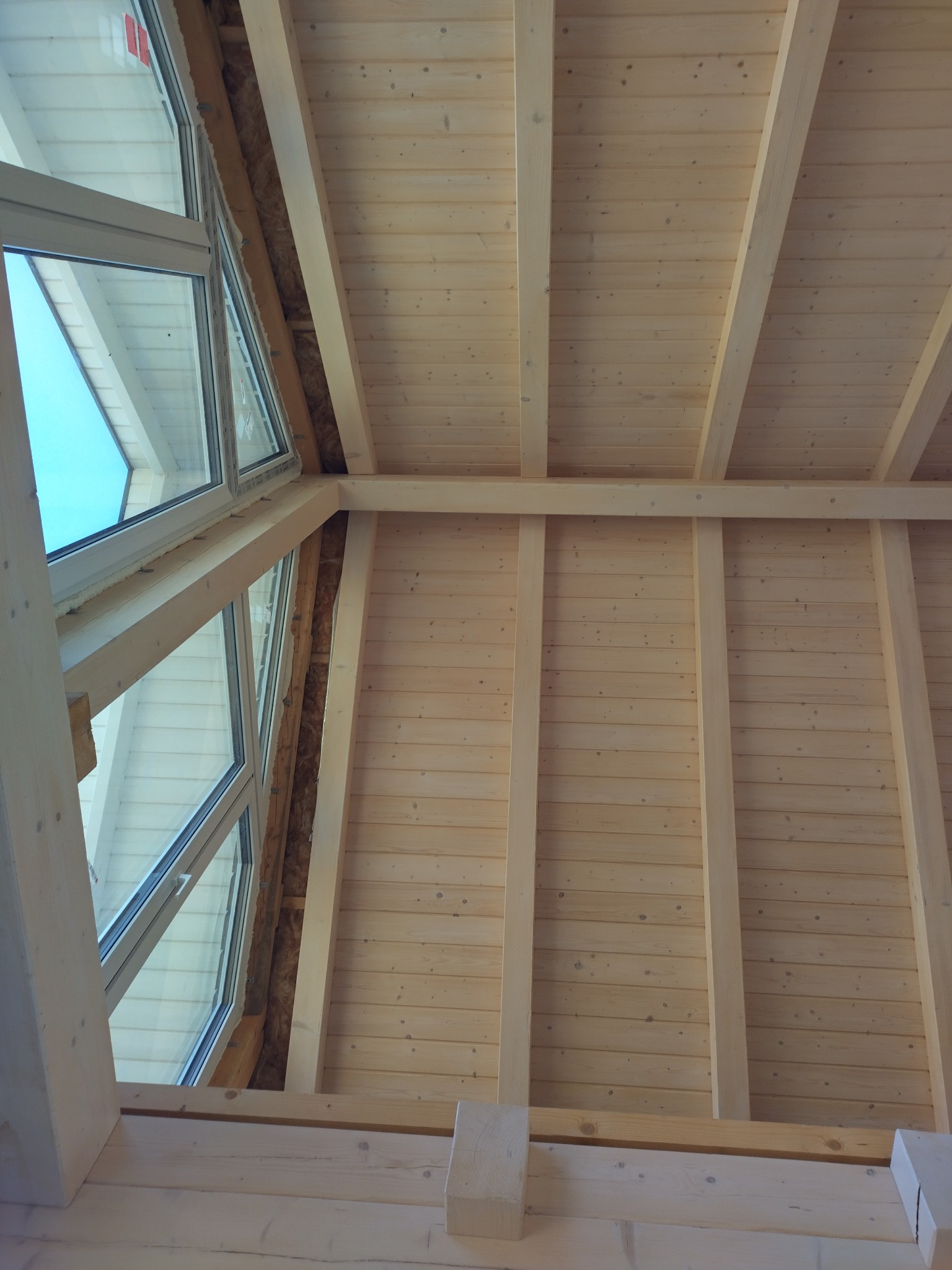 Wnętrze w trakcie budowy: surowe drewno, widoczne elementy konstrukcyjne ścian i stropu, okno dachowe z widokiem na błękitne niebo. Widoczne ocieplenie wokół okna.