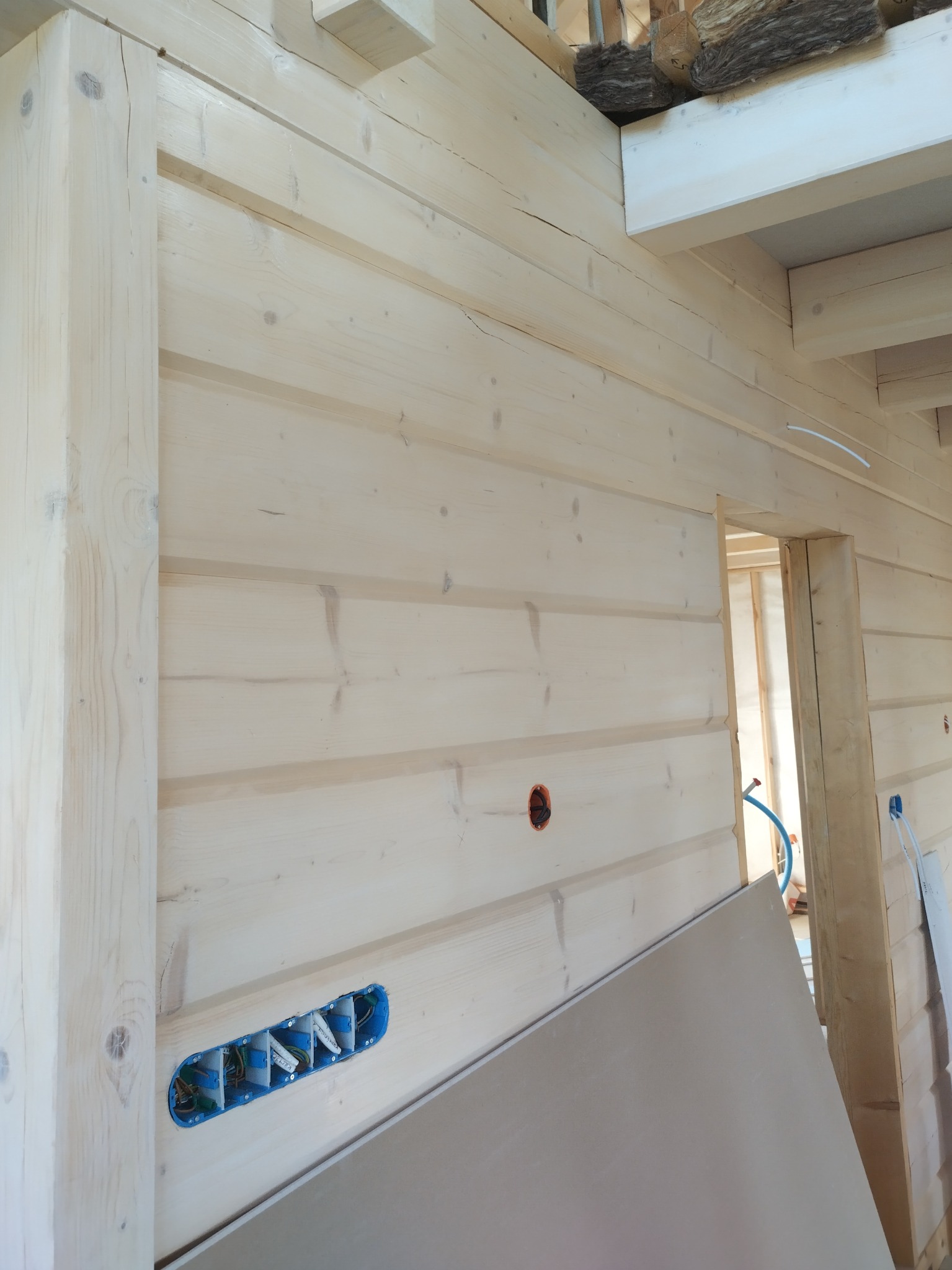 Wnętrze w trakcie wykańczania, ściana z jasnych bali z widocznymi puszkami elektrycznymi i fragmentem płyty gipsowo-kartonowej.