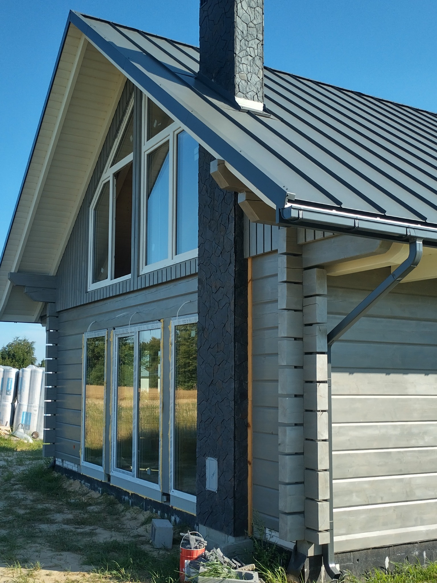 Szary dom z bali z ciemnoszarą blachodachówką i kamiennym kominem, widoczne okna z białymi ramami i rynna, w tle błękitne niebo i materiały budowlane.