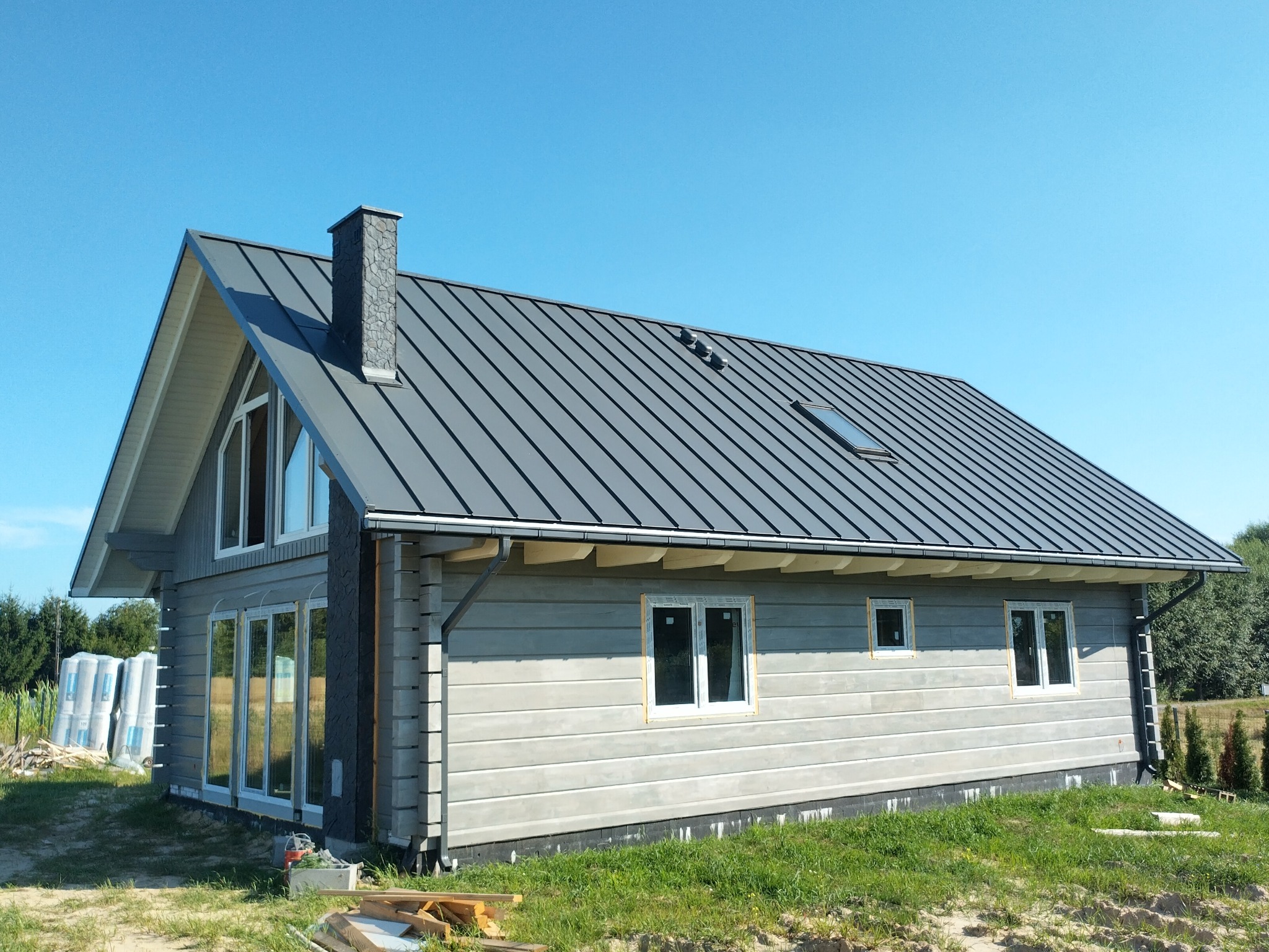 Szary dom z bali z ciemnoszarą blachodachówką i kamiennym kominem, okna jeszcze bez wykończenia, w tle błękitne niebo.