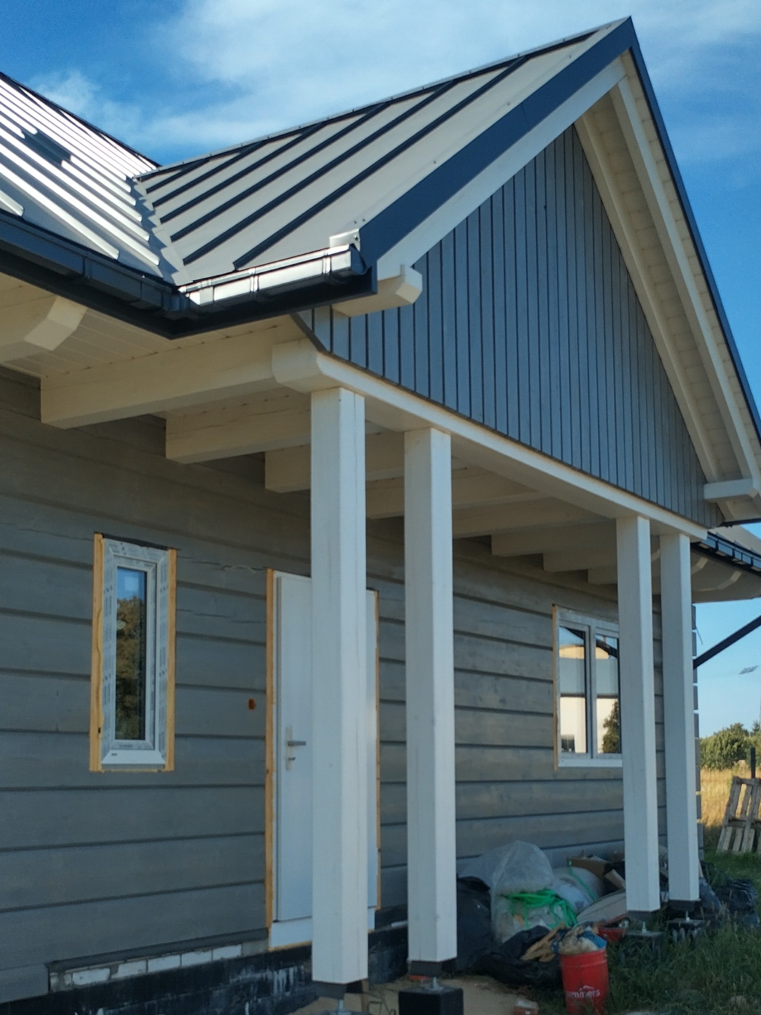 Szary dom w trakcie budowy z widoczną konstrukcją dachu, białe kolumny podtrzymujące zadaszenie wejścia, niezamontowane okno i materiały budowlane na pierwszym planie.