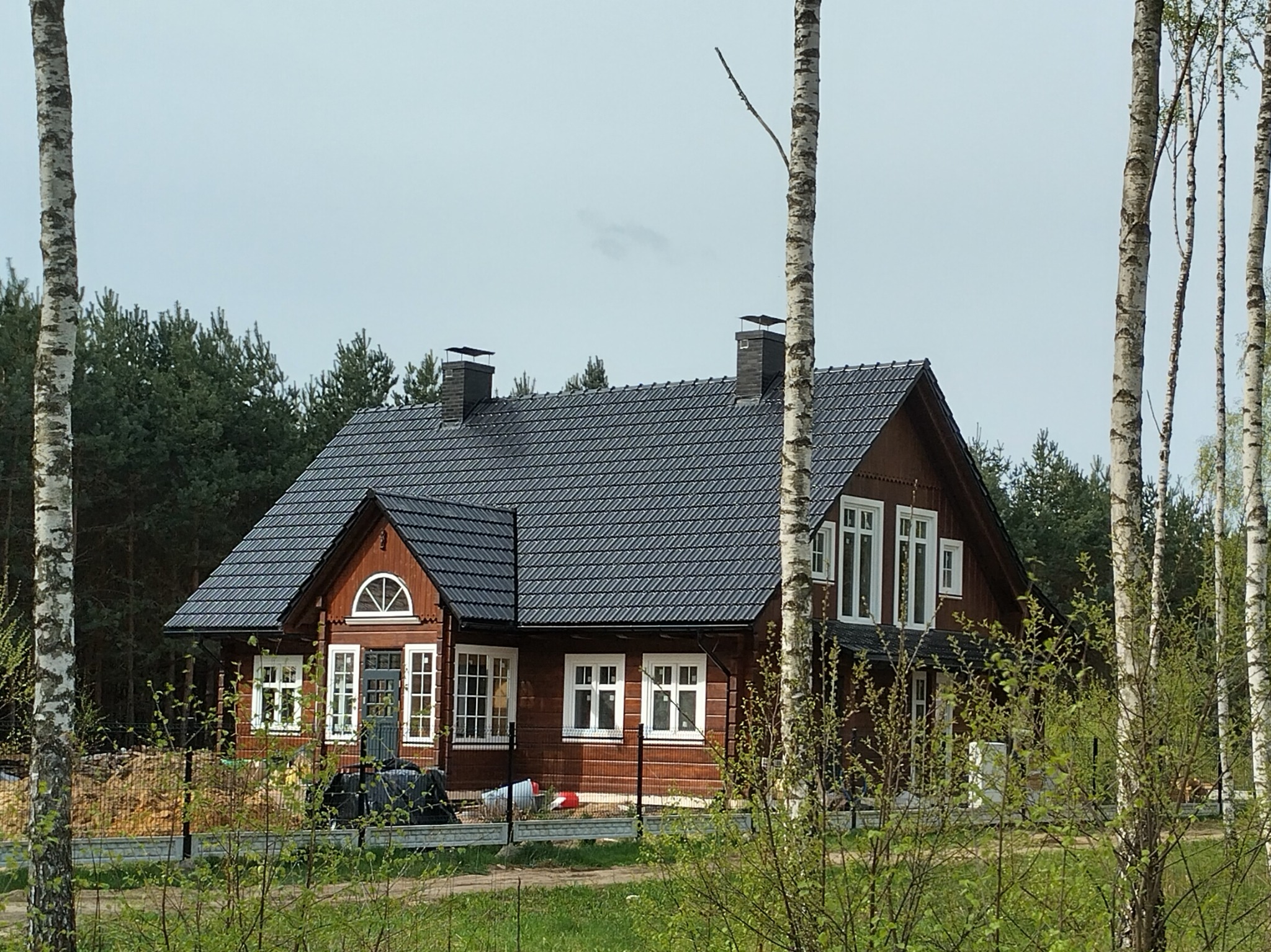 Gotowy, brązowy dom z ciemną dachówką, widoczny zza drzew brzozowych i ogrodzenia, z fragmentem placu budowy w tle.