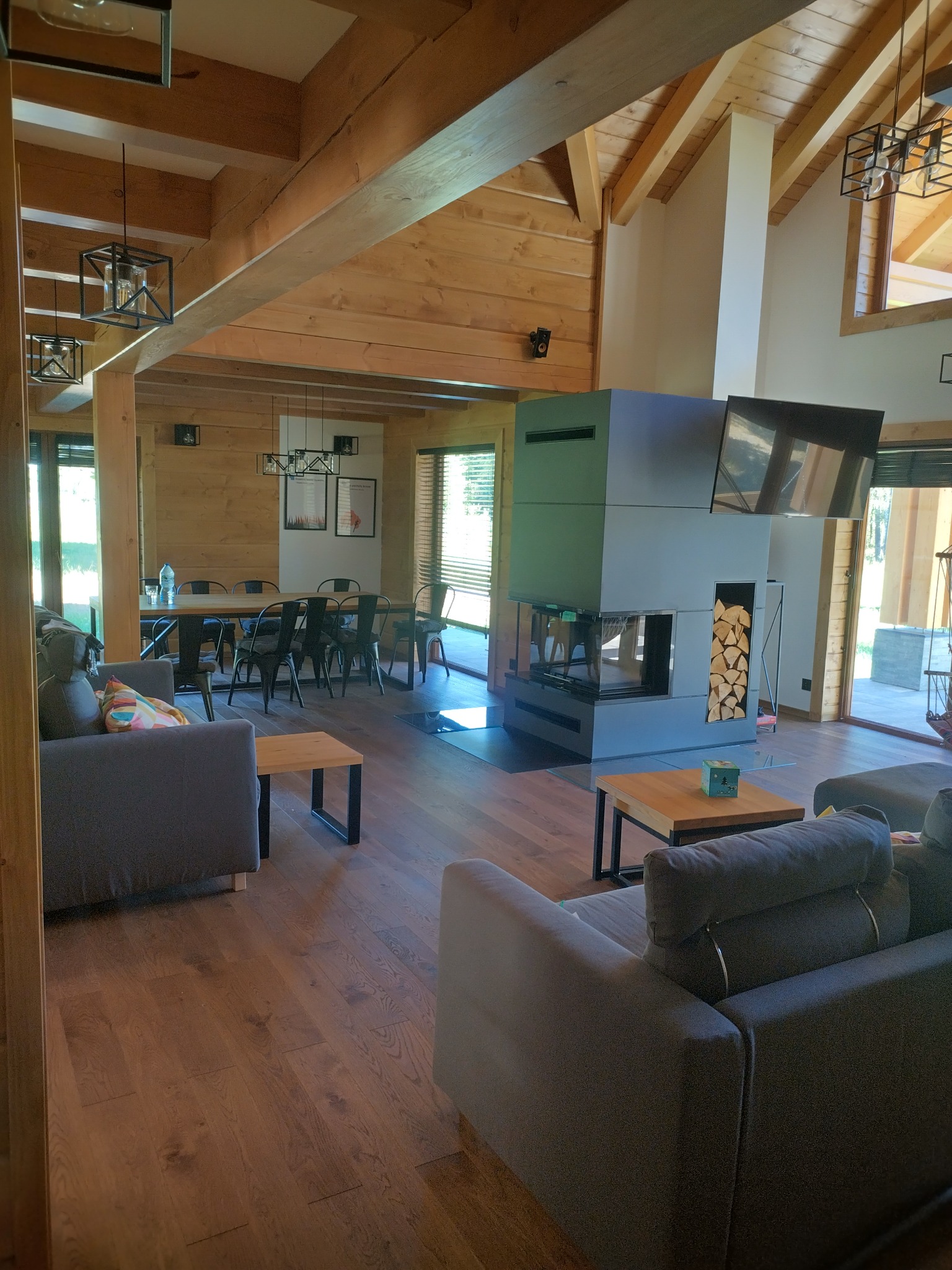 Wnętrze domu z bali z drewnianą konstrukcją stropu, nowoczesnym kominkiem z wnęką na drewno i szarymi sofami.