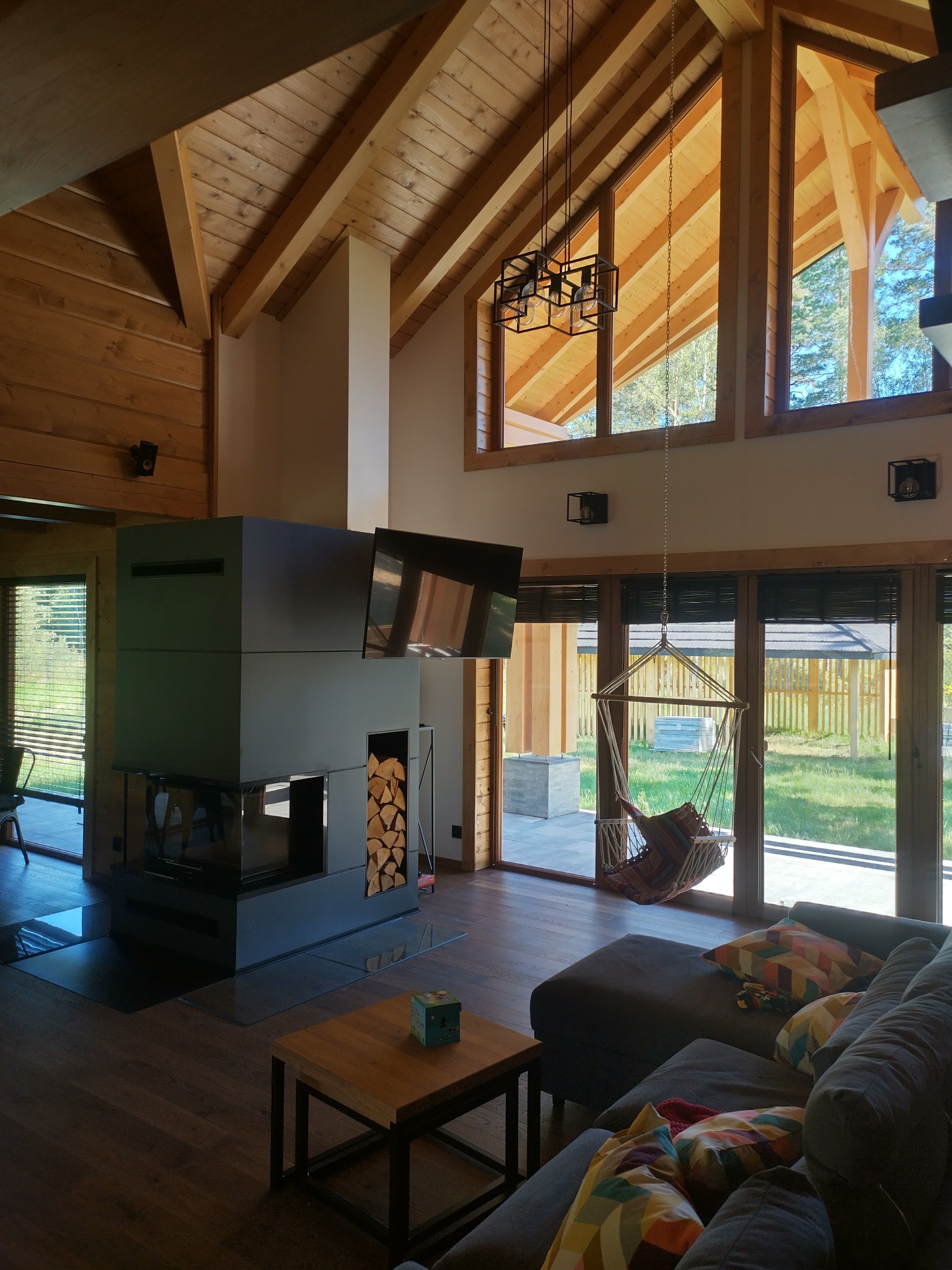 Jasne wnętrze domu z drewnianą konstrukcją dachu, nowoczesnym kominkiem z zapasem drewna, wiszącym fotelem i dużymi oknami z widokiem na zewnątrz.