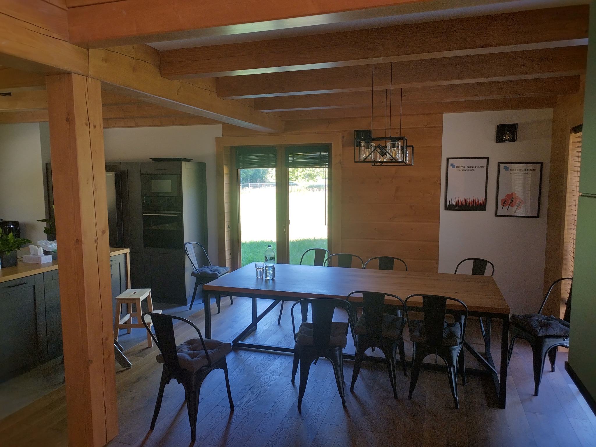 Wnętrze domu z bali: drewniane belki stropowe, duży stół z metalowymi nogami, czarne krzesła z poduszkami, lampa wisząca w stylu industrialnym, okno z widokiem na zieleń.