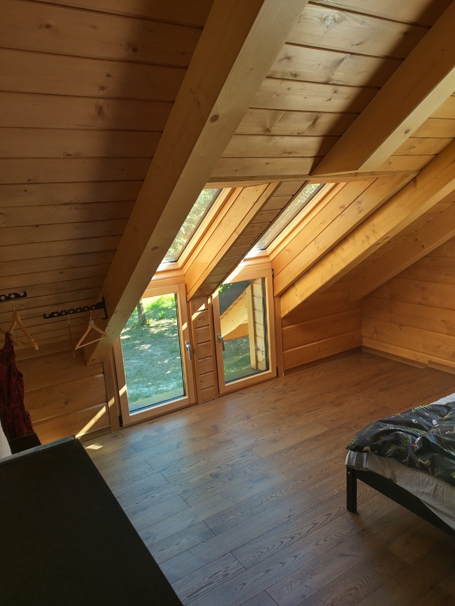 Wnętrze poddasza z drewnianą boazerią, oknem dachowym i widokiem na zieleń, z fragmentem łóżka i wieszakami na ubrania.