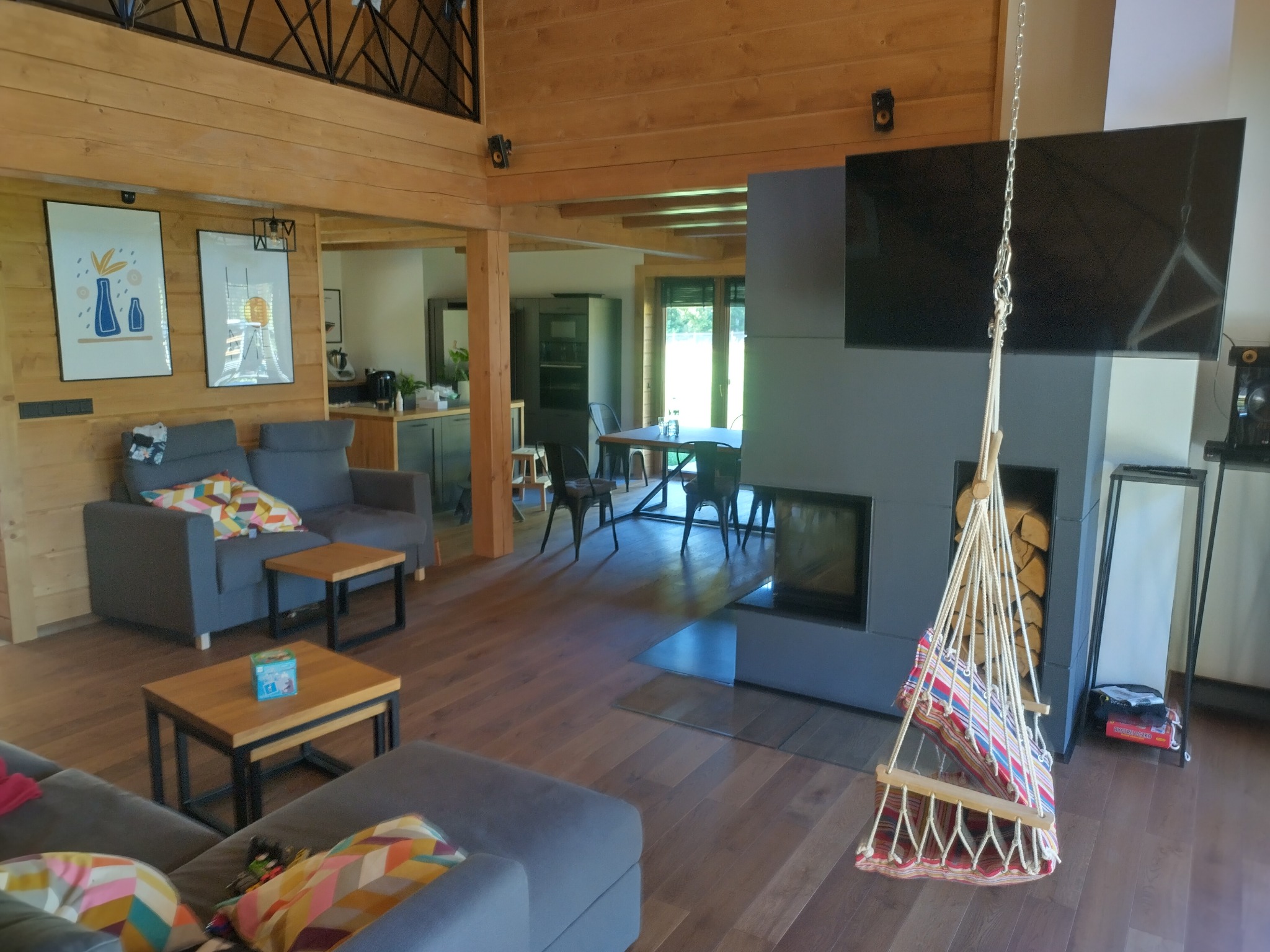 Jasne wnętrze domu z bali z drewnianymi ścianami, szarymi sofami, wiszącym fotelem hamakowym i nowoczesnym kominkiem z telewizorem