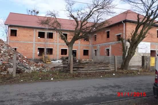 Budynek dwukondygnacyjny w stanie surowym otwartym z czerwonej cegły, widoczny stos desek i gruzu, drzewo bez liści na pierwszym planie, data 31/1/2015 w prawym dolnym rogu.