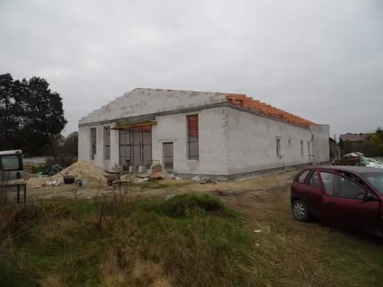 Niedokończony parterowy budynek z białych bloczków, z częściowo ułożoną dachówką, widoczny stos piasku i czerwony samochód osobowy na pierwszym planie.