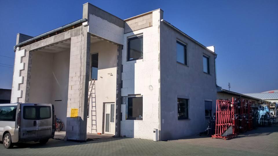 Budynek w trakcie ocieplania styropianem, widoczna drabina oparta o ścianę, zaparkowany srebrny bus i stojaki z szybami w tle.