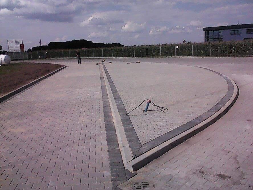 Ułożona kostka brukowa na placu, z widocznym odpływem liniowym i krawężnikiem, osoba w tle, budynek w oddali.