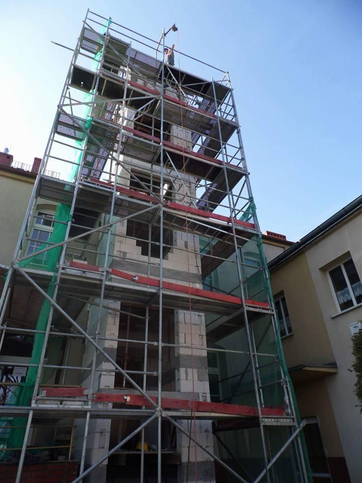 Wznoszenie wielopiętrowej konstrukcji z bloczków na zewnątrz budynku, otoczonej metalowym rusztowaniem z siatką zabezpieczającą, widok z dołu.