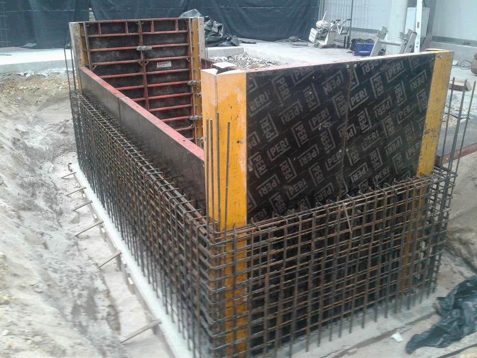 Szczegółowy widok zbrojenia i szalunków fundamentów, z widocznymi elementami drewnianymi i stalowymi, na placu budowy.