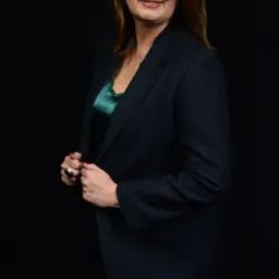 Portret kobiety w okularach i ciemnym garniturze na czarnym tle, z delikatnym uśmiechem i widocznym pierścionkiem na palcu.