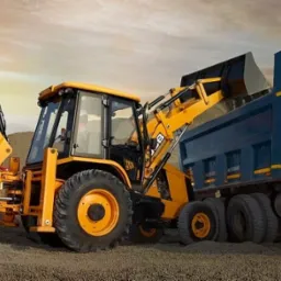 Żółta koparko-ładowarka JCB ładuje ziemię do niebieskiej wywrotki na tle piaszczystego terenu i zachodzącego słońca.