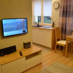 Jasne wnętrze pokoju z aneksem kuchennym: telewizor na ścianie, białe szafki kuchenne z drewnianym blatem, stół z krzesłem i dekoracje.