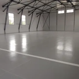 Jasnoszara, błyszcząca podłoga w hali sportowej z białymi liniami wyznaczającymi pole gry, widoczne okna i metalowa konstrukcja dachu.