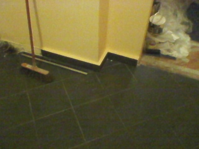 Podłoga z ciemnych płytek po remoncie, widoczna szczotka i listwa przypodłogowa, ślady farby na ścianie i podłodze.