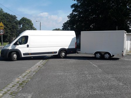 Biały zestaw transportowy: Ducato z przyczepą kontenerową DMC 5t, zaparkowany na parkingu, znak ograniczenia prędkości w tle.