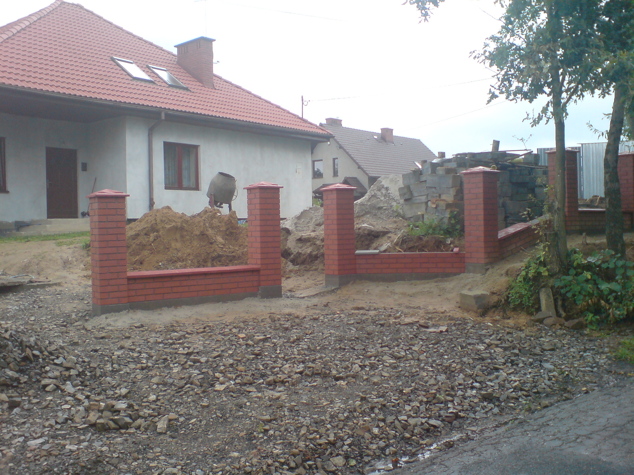 Wznoszenie ogrodzenia z czerwonej cegły klinkierowej na nierównym terenie, widoczna sterta ziemi, gruz i materiały budowlane, w tle dom z czerwonym dachem i mieszalnik do betonu.