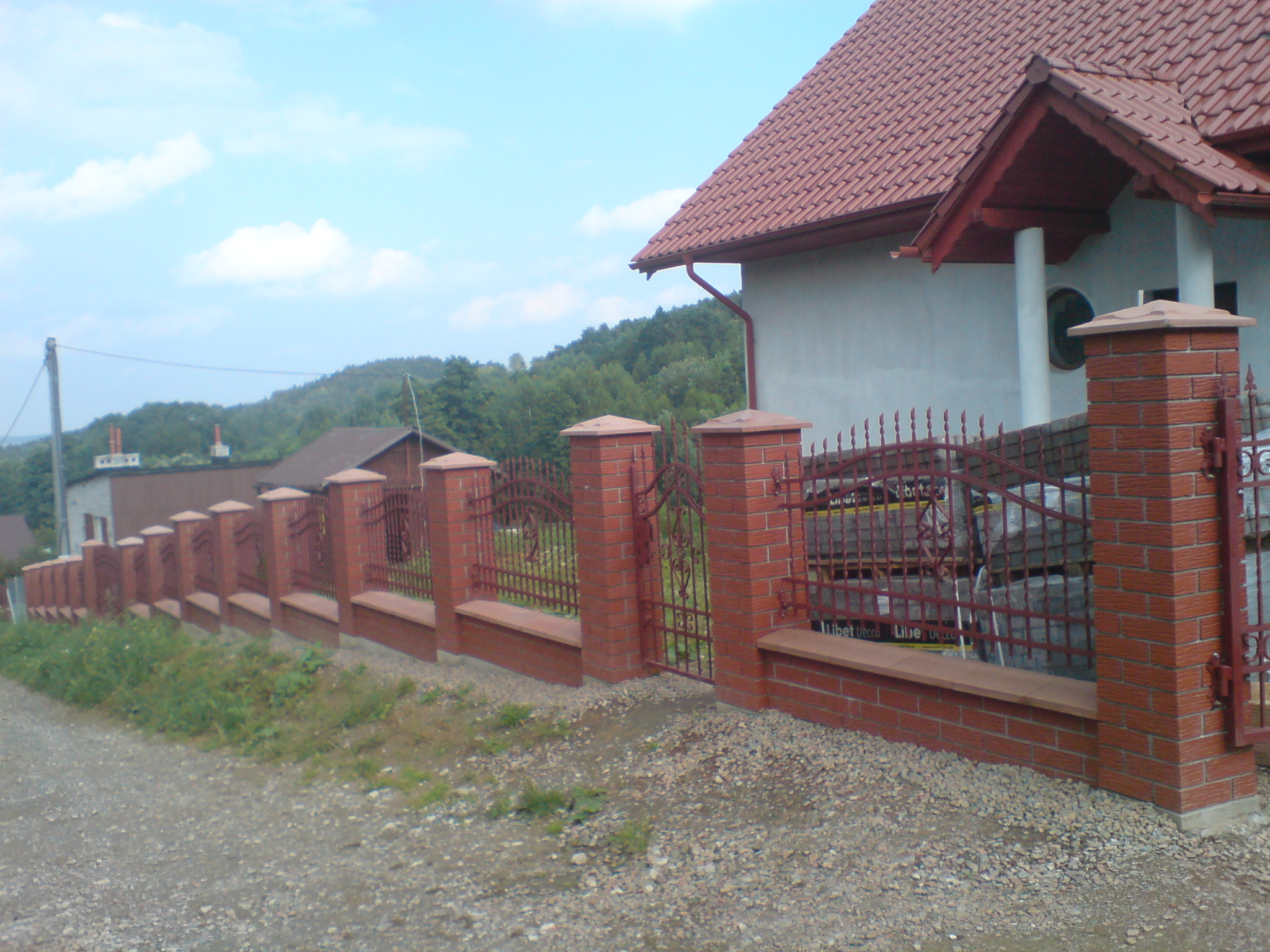 Czerwono-ceglany, częściowo ażurowy płot z elementami kutymi, z podmurówką i słupkami, otaczający posesję z domem z czerwoną dachówką w tle, widok z drogi szutrowej.