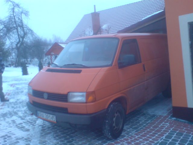Pomarańczowy bus VW Transporter częściowo wjeżdża do garażu, zimowa sceneria z śniegiem w tle.