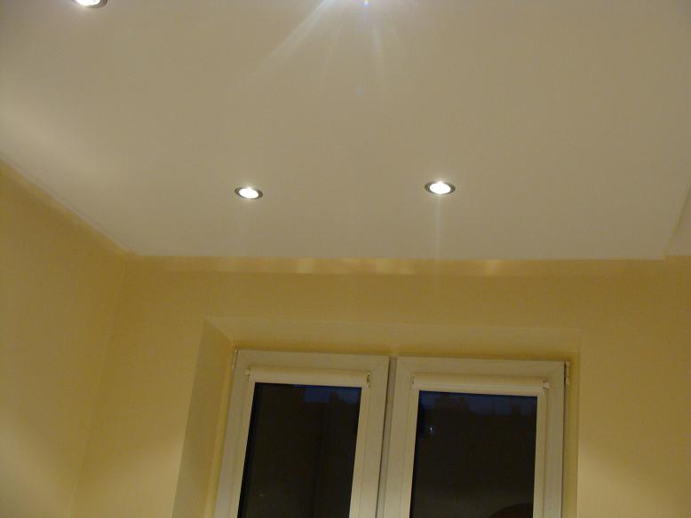 Biały, matowy sufit z trzema punktowymi lampami LED, widok na fragment ściany pomalowanej na żółto i okno z roletą.