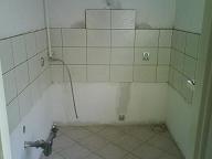 Niedokończona łazienka z częściowo położonymi jasnymi płytkami na ścianach i podłodze, widoczne rury i niedokończone fugi, surowy stan przed wykończeniem.