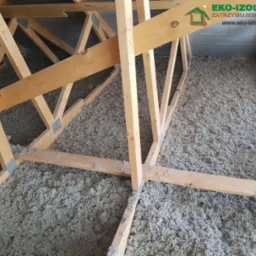 Wypełnienie izolacyjne z wełny celulozowej w przestrzeni wiązarów drewnianych dachu, widoczne metalowe łączniki belek. Na górze widoczne logo firmy Eko-Izolacje.