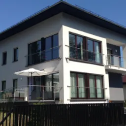 Nowoczesny, dwukondygnacyjny dom z balkonami i szklanymi balustradami, widziany zza czarnego, metalowego ogrodzenia. Biało-szara elewacja i duże okna.