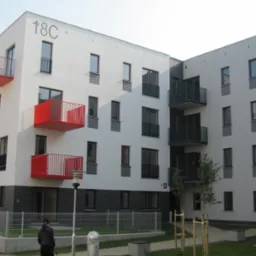 Nowoczesny budynek wielorodzinny z białymi elewacjami, czerwonymi balkonami i numerami '18C' i '18D'. Widoczne okna, lampa i fragment chodnika z osobą.