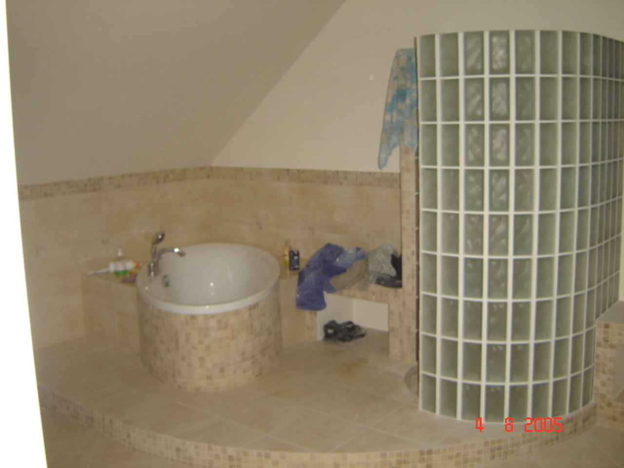 Zaokrąglona wanna obudowana mozaiką w łazience na poddaszu, z kabiną prysznicową z luksferów i beżowymi płytkami podłogowymi.