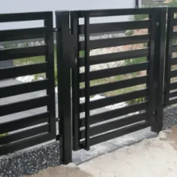 Nowoczesna, czarna brama palisadowa z widocznym zamkiem, zamontowana na betonowej podstawie, z widokiem na ogród z białymi kamieniami.