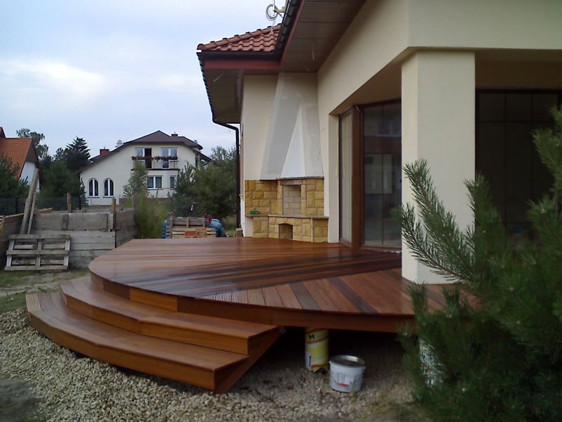 Drewniany taras z półokrągłymi schodami, widoczne puszki z farbą pod spodem, dom z kamiennym kominkiem w tle.