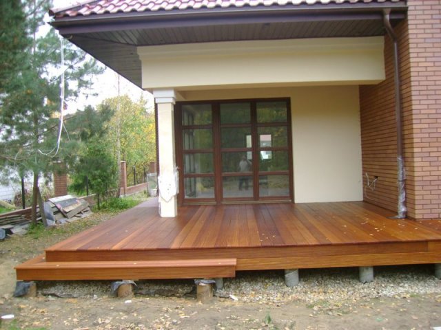 Nowoczesny taras z drewna egzotycznego z niskimi schodkami, widok na dom z brązowymi oknami i ceglaną ścianą, w tle zieleń.