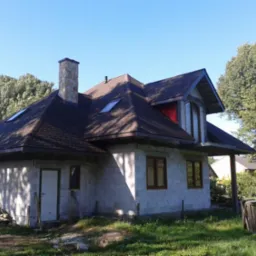 Surowy dom z keramzytu w Pawłowie z brązowym dachem i kamiennym kominem. Widoczne okna i drzwi. Budynek w otoczeniu zieleni i drzew.