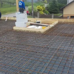 Zbrojenie płyty fundamentowej z przygotowanym otworem na komin, widoczni pracownicy w tle.