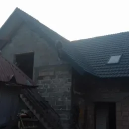 Budynek w trakcie budowy z zamontowanym, ciemnym dachem z dwoma oknami dachowymi, widoczne fragmenty muru z pustaków, drewniana wieżyczka z boku.