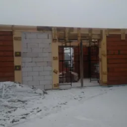 Wznoszenie ścian parterowego budynku z czerwonej cegły i białego betonu komórkowego na zimowym placu budowy, widoczne drewniane szalunki i zbrojenia.