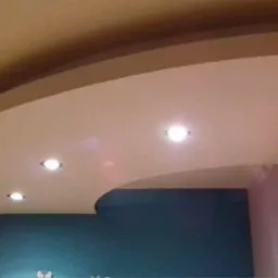 Podwieszany sufit z gładzi gipsowej, o nieregularnym kształcie z trzema punktami świetlnymi, na tle ściany pomalowanej na ciemny turkusowy kolor.