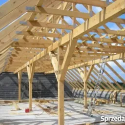 Drewniana konstrukcja dachu w trakcie budowy, widoczne krokwie, słupy i płatwie połączone metalowymi złączkami, drabina oparta o konstrukcję, w tle mur z bloczków betonowych i błękitne niebo.