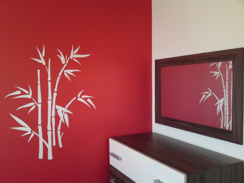 Czerwona ściana z białym motywem bambusa, odbitym również w lustrze z ciemną ramą, nad biało-brązową komodą z szufladami.