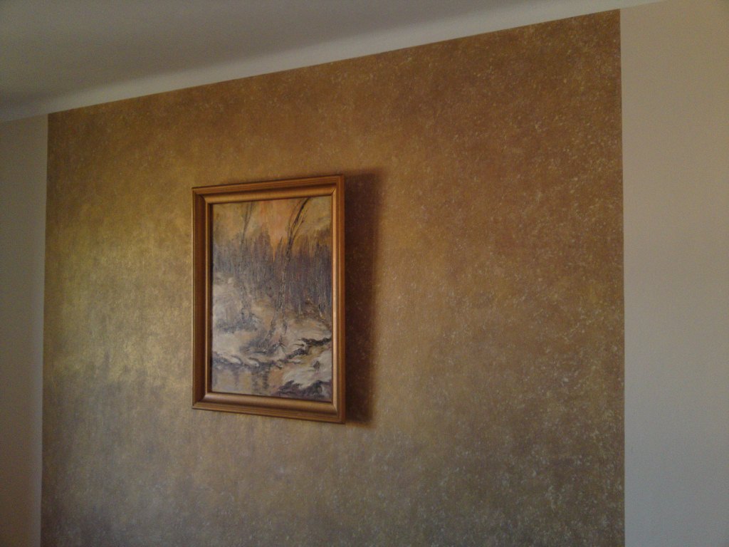 Ściana pokryta tynkiem dekoracyjnym w odcieniach złota i brązu, z zawieszonym obrazem w złotej ramie.