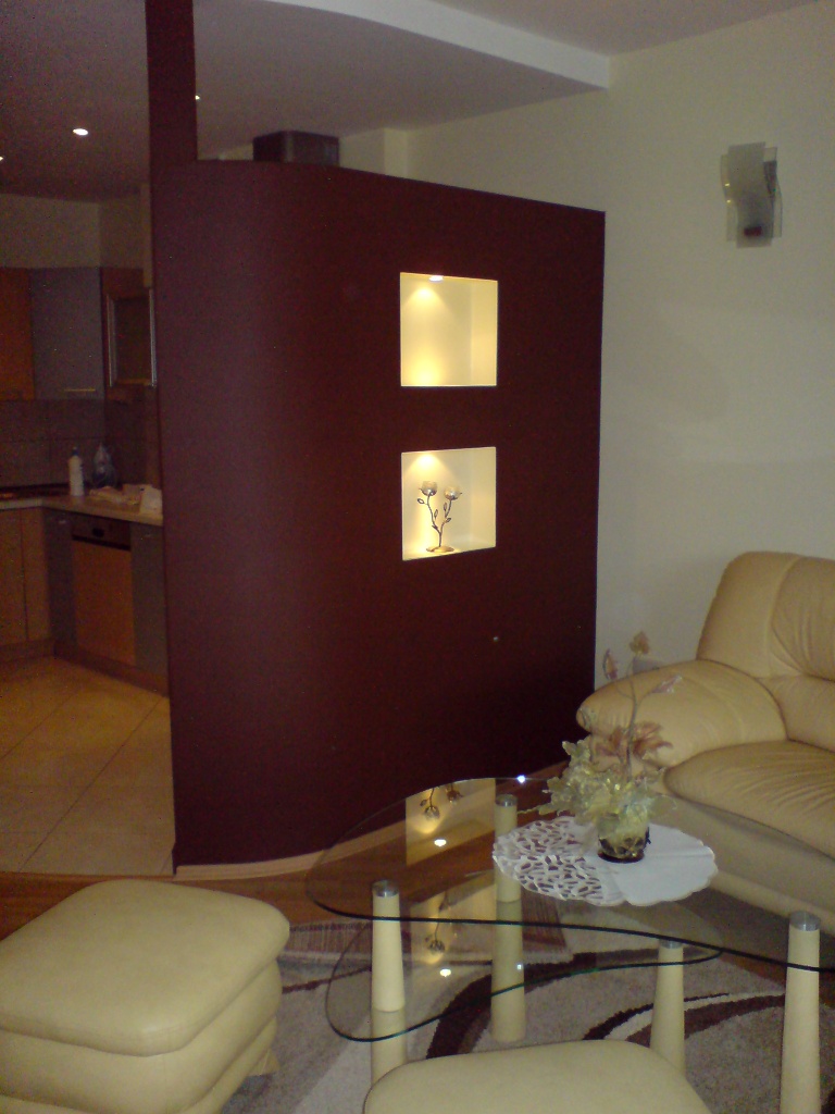 Ciemnoczerwona ścianka działowa z podświetlanymi niszami w salonie, widok na kuchnię w tle.